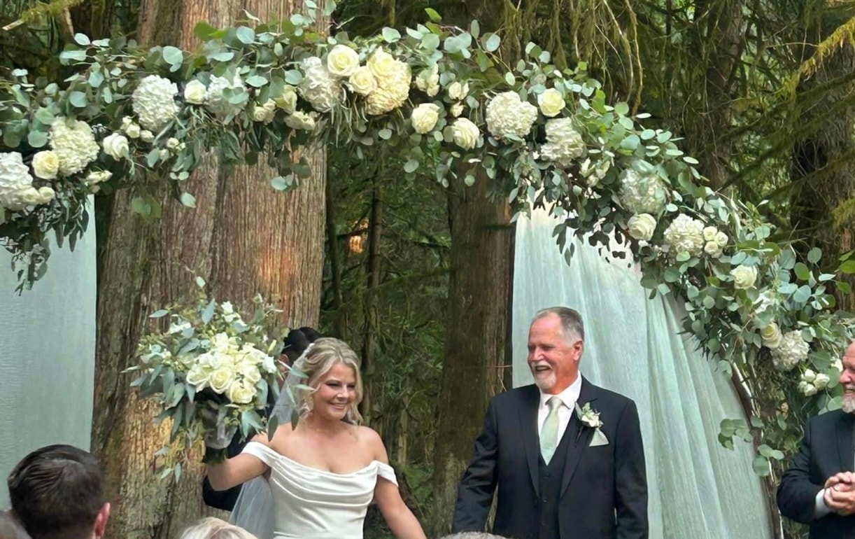 Bride and father walking under a lush white floral arch in the forest
